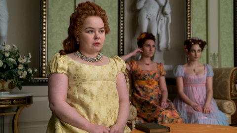 Nicola Coughlan as Penelope Featherington sits at a table with Bessie Carter as Prudence Featherington and Harriet Cains as Phillipa Featherington in the background in episode 10 of Bridgerton
