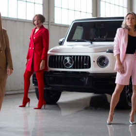 Three women posing in front of a Mercedes-Benz wearing Pink Tartan power suits