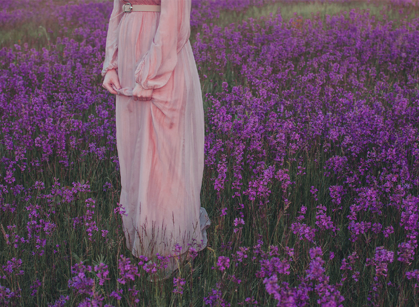A woman wearing a pink, long-sleeved ankle length dress shown from the waist down and standing in a field of flowers