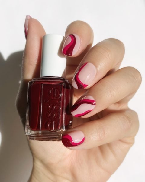 a hand with abstract nail polish designs holding a berry coloured bottle of Essie nail polish