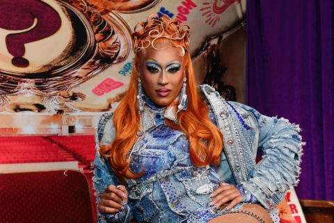 drag queen priyanka poses sitting down with orange hair and jean jacket and jean bodysuit