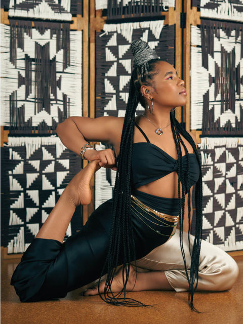 woman holds a yoga pose with long grey braids, back cutout top and black pants