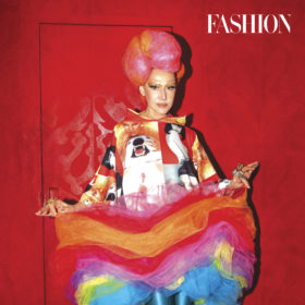 susanne bartsch wears a pink wig, big rainbow tulle skirt and sweater with photos on it against a red backdrop