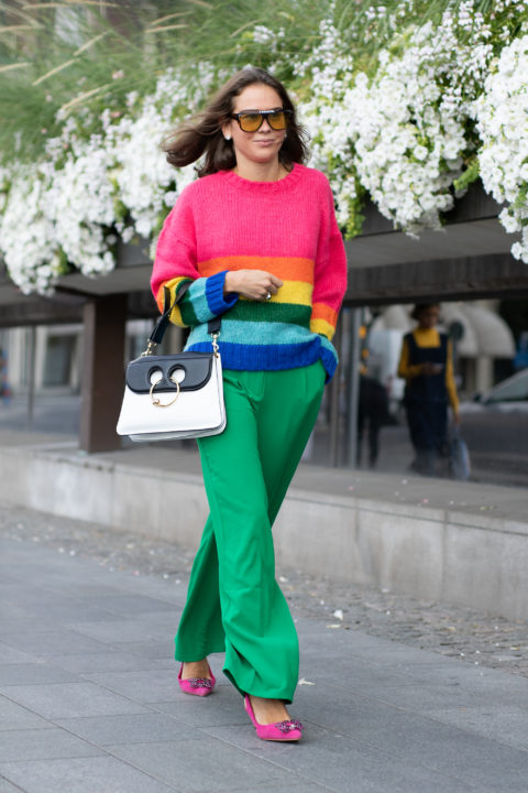 woman in a rainbow sweater with green pants and a black and white purse