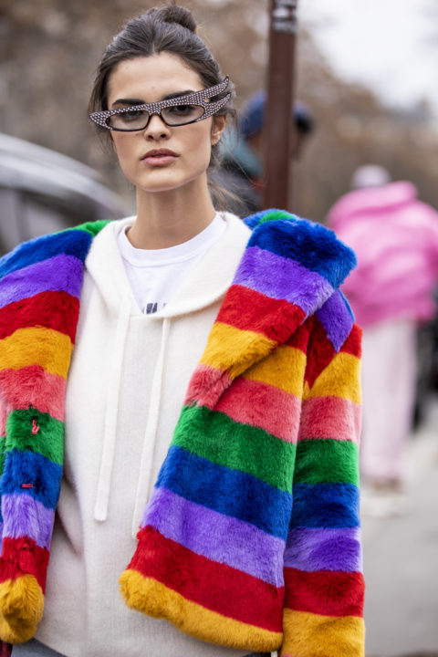 close-up of a woman in a white sweater with a fluffy rainbow jacket and large glasses