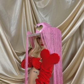 a model wears a pink cowboy hat with a cutout heart at the front and long strings. They also have on a red bra and underwear with long oversized red gloves 