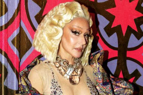 susanne bartsch wears a blonde wig, big chunky necklace, sheer top and metallic jacket against a purple and red graphic backdrop