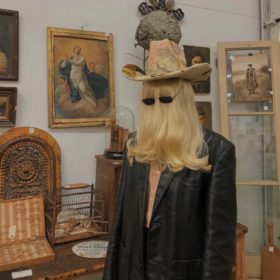 model wears a blonde wig that obscures their face and sunglasses with a vintage cowboy hat and black blazer. they are surrounded by antiques