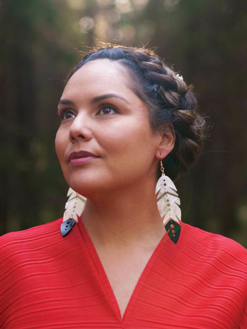 person wears indi city eagle feather earrings with hair braded out of their face and red shirt. they are in nature