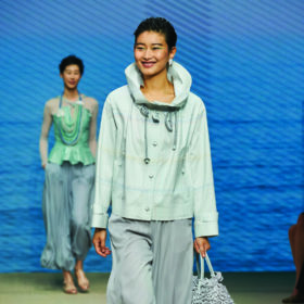 a smiling model in a blue sweater and pants with a shaved head walks the runway