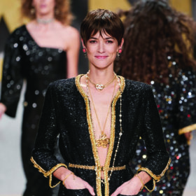 smiling models walk down the chanel spring runway. the model in the forefront has a pixie cut and wears a black and gold blazer with nothing underneath and lots of jewellery