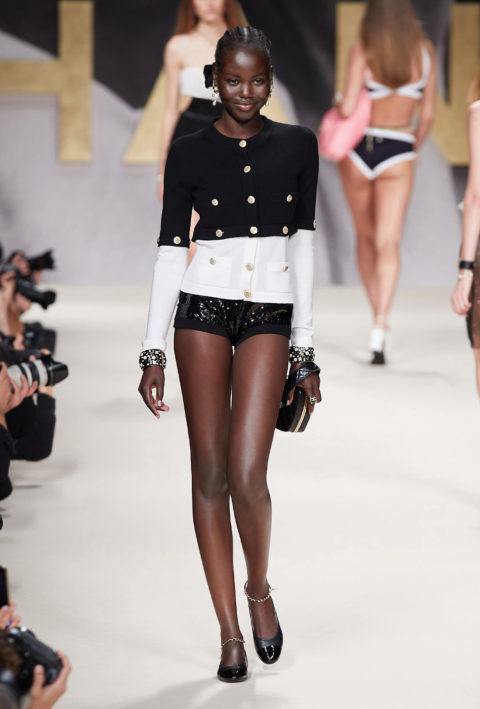 a smiling model walks down the chanel spring runway in a black and white cardigan and short black shorts