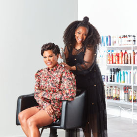Janet Jackson and Marci Len pose inside one of the most popular curly hair salons in toronto, JouJou Hair studio. one woman sits on a chair while the other stands behind her