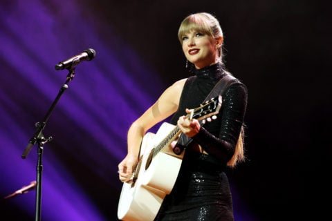 Taylor Swift wears a black turtleneck dress and her hair back while playing guitar on stage