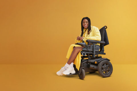A photo of a smiling young woman with disability sitting in wheelchair against yellow background
