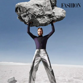 Myles Sexton holds a large rock above his head while wearing silver pants and a black and dark purple turtleneck